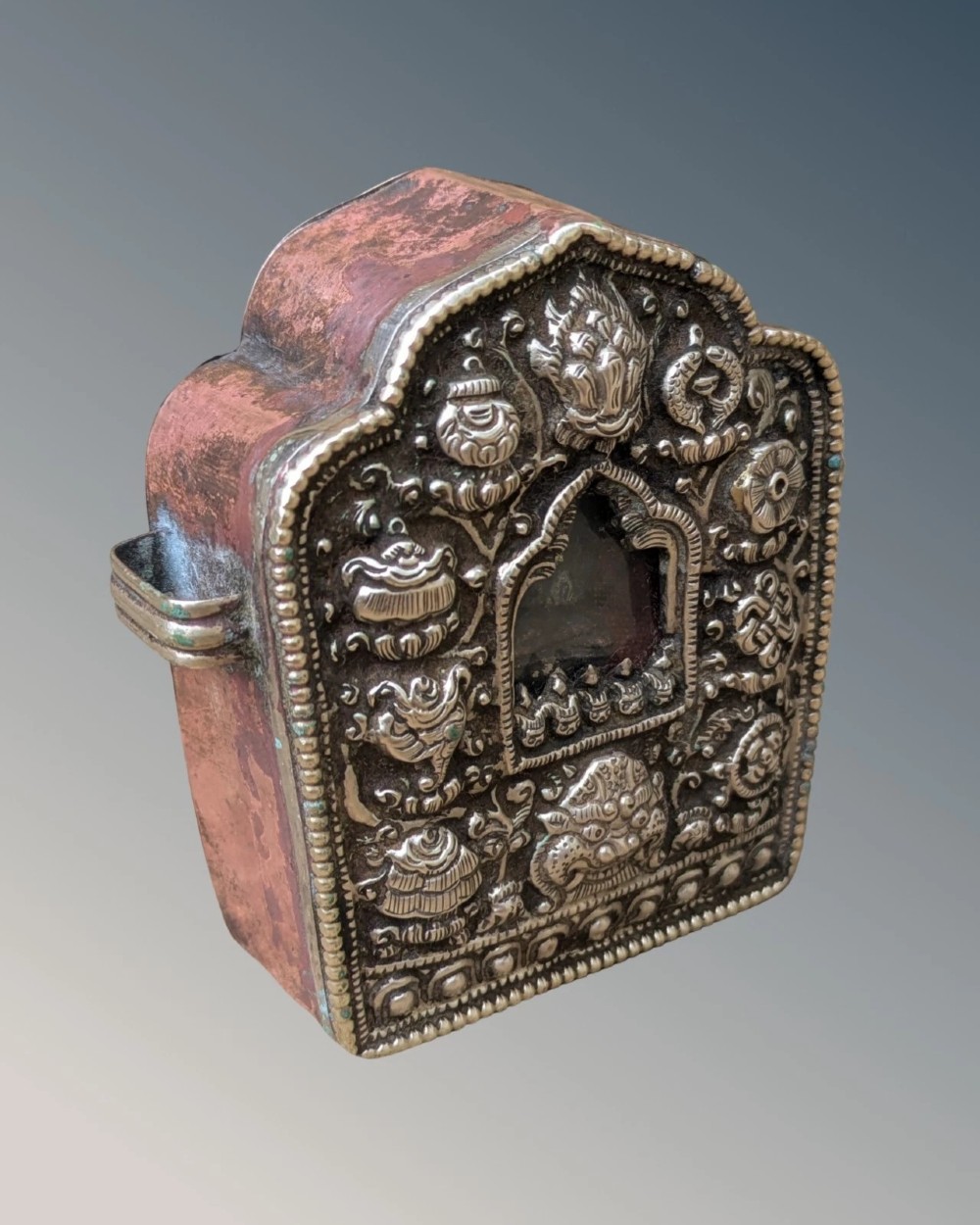 An arched Tibetan Gau prayer shrine featuring an intricately embossed silver metal front panel mounted on a copper casing with side loops. View III