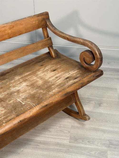 fig 1. A long antique wooden rocking settle featuring open back slats, curved scroll armrests, and a solid plank seat resting on wooden rocking runners.