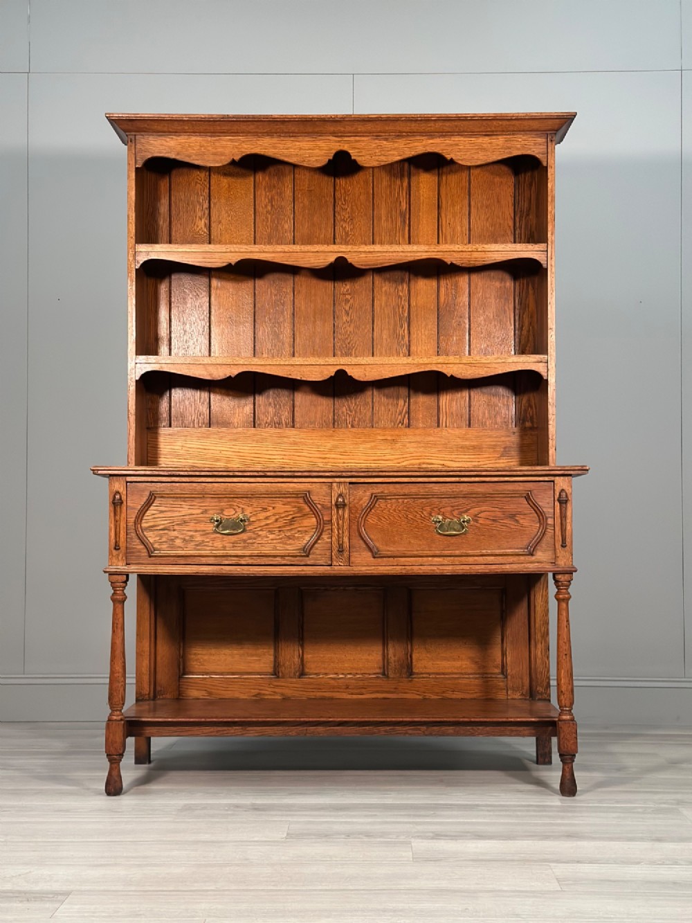 An antique oak Welsh dresser featuring a three-tier open plate rack above two moulded drawers with brass handles and a lower open potboard shelf supported by turned front legs.