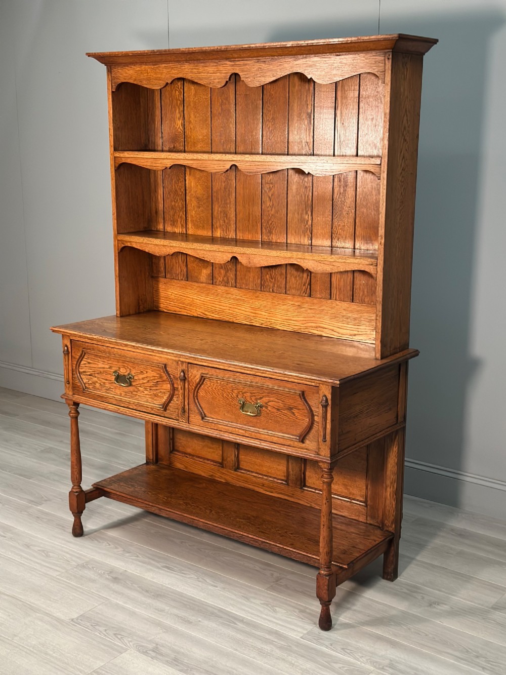 An antique oak Welsh dresser featuring a three-tier open plate rack above two moulded drawers with brass handles and a lower open potboard shelf supported by turned front legs. View II