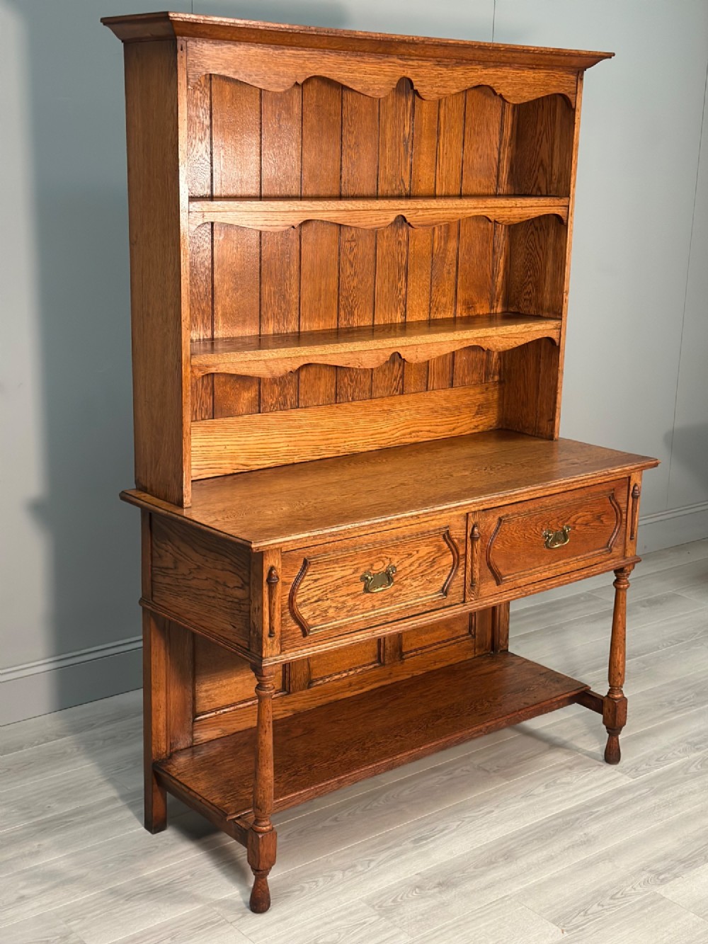 An antique oak Welsh dresser featuring a three-tier open plate rack above two moulded drawers with brass handles and a lower open potboard shelf supported by turned front legs. View IV