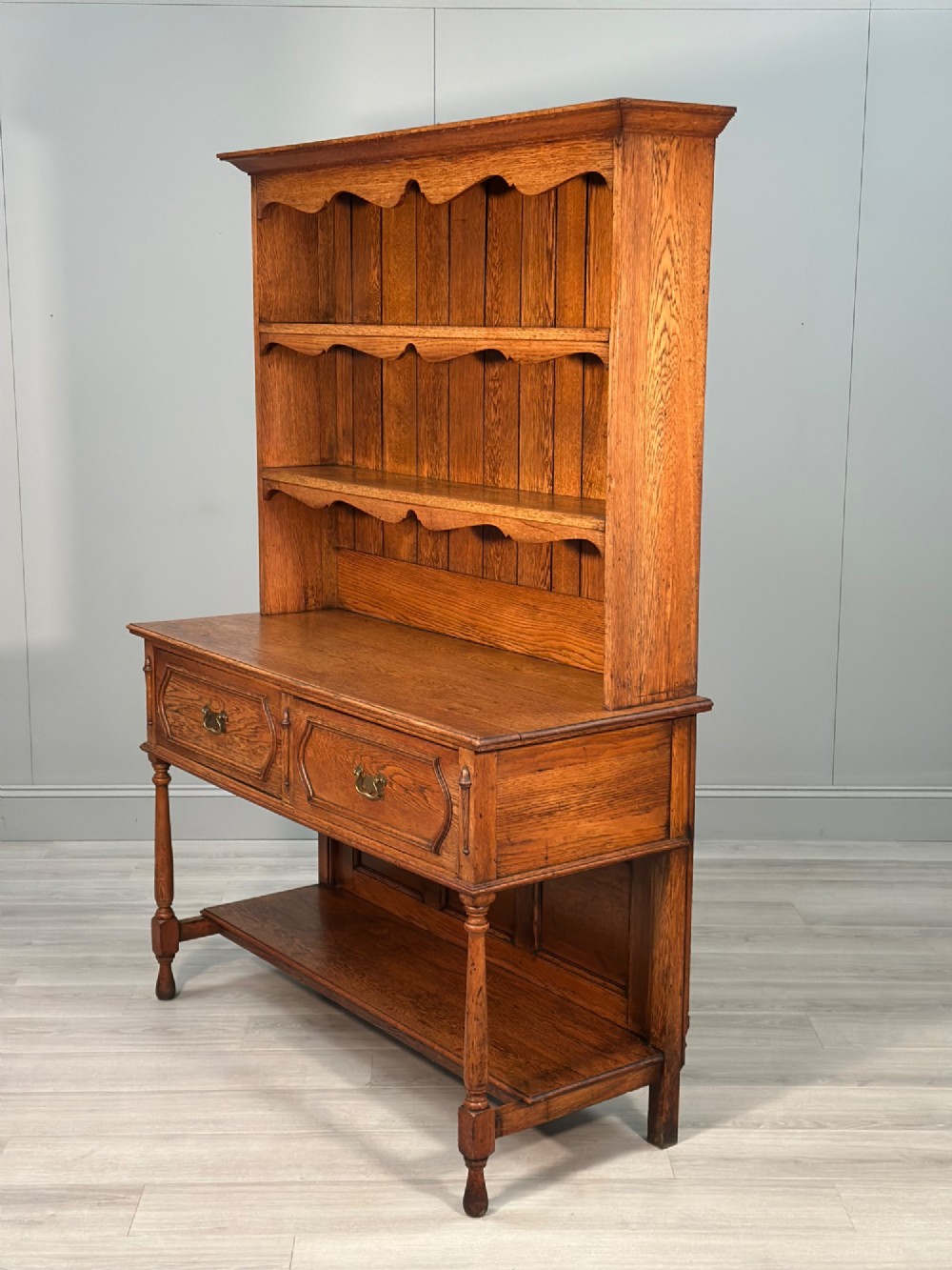An antique oak Welsh dresser featuring a three-tier open plate rack above two moulded drawers with brass handles and a lower open potboard shelf supported by turned front legs. View XII