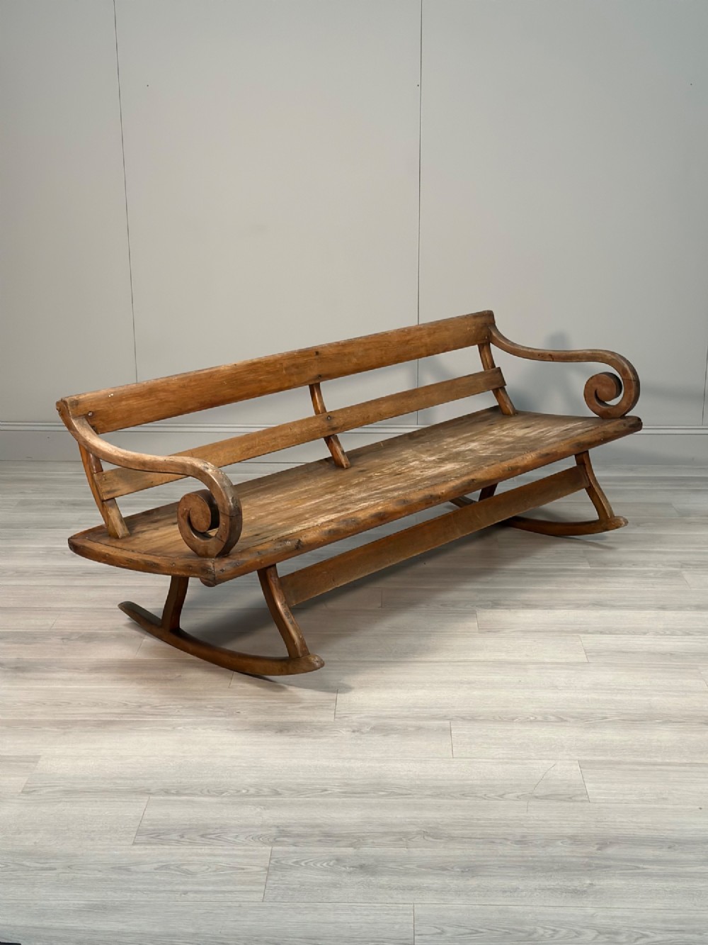 A long antique wooden rocking settle featuring open back slats, curved scroll armrests, and a solid plank seat resting on wooden rocking runners.