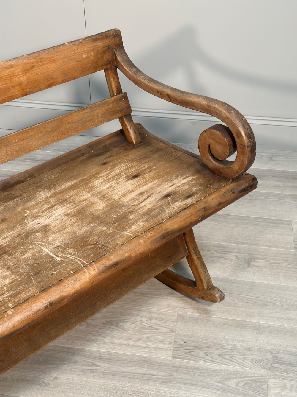 A long antique wooden rocking settle featuring open back slats, curved scroll armrests, and a solid plank seat resting on wooden rocking runners. View II