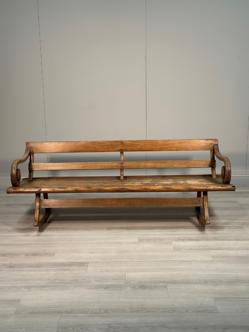 A long antique wooden rocking settle featuring open back slats, curved scroll armrests, and a solid plank seat resting on wooden rocking runners. View IV