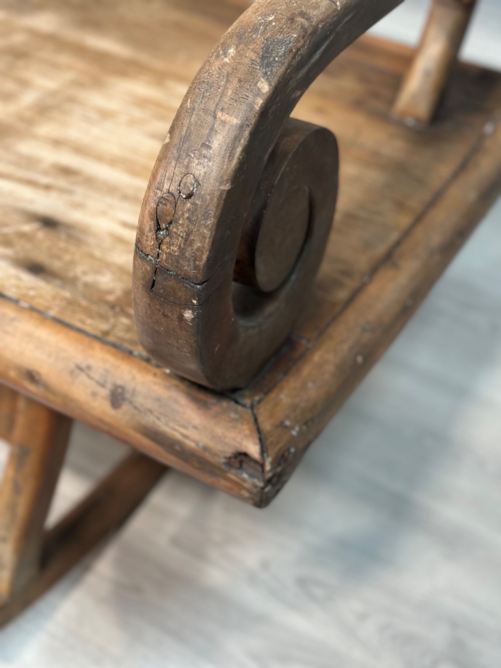 A long antique wooden rocking settle featuring open back slats, curved scroll armrests, and a solid plank seat resting on wooden rocking runners. View V