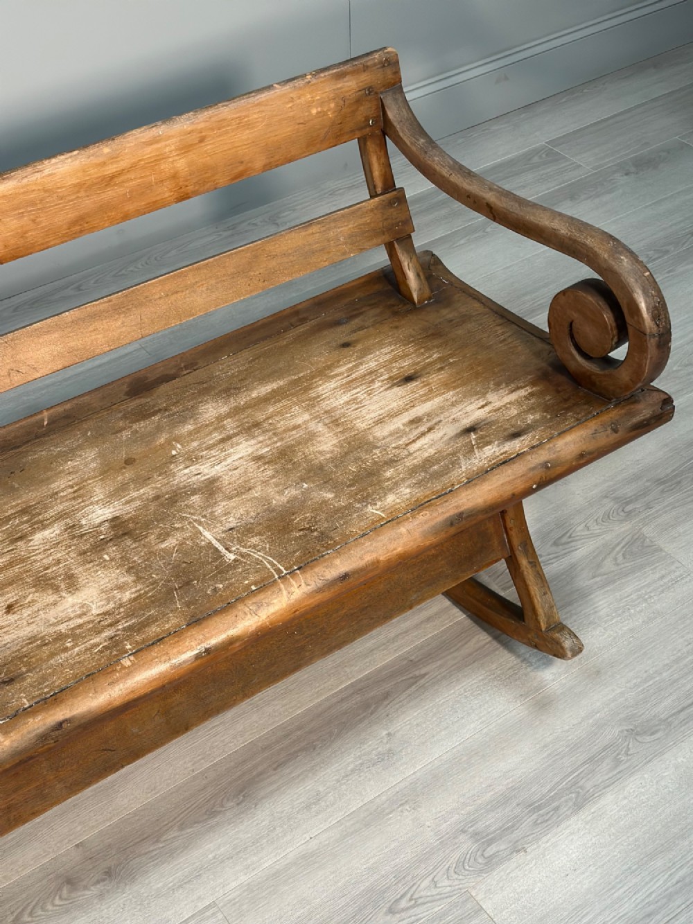 A long antique wooden rocking settle featuring open back slats, curved scroll armrests, and a solid plank seat resting on wooden rocking runners. View VI