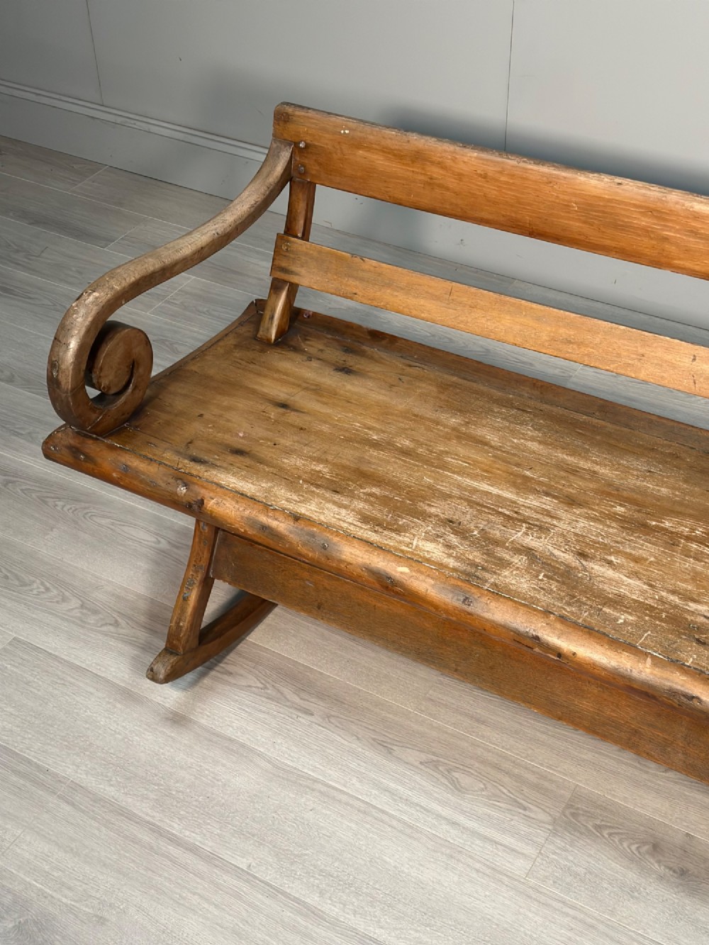 A long antique wooden rocking settle featuring open back slats, curved scroll armrests, and a solid plank seat resting on wooden rocking runners. View VII