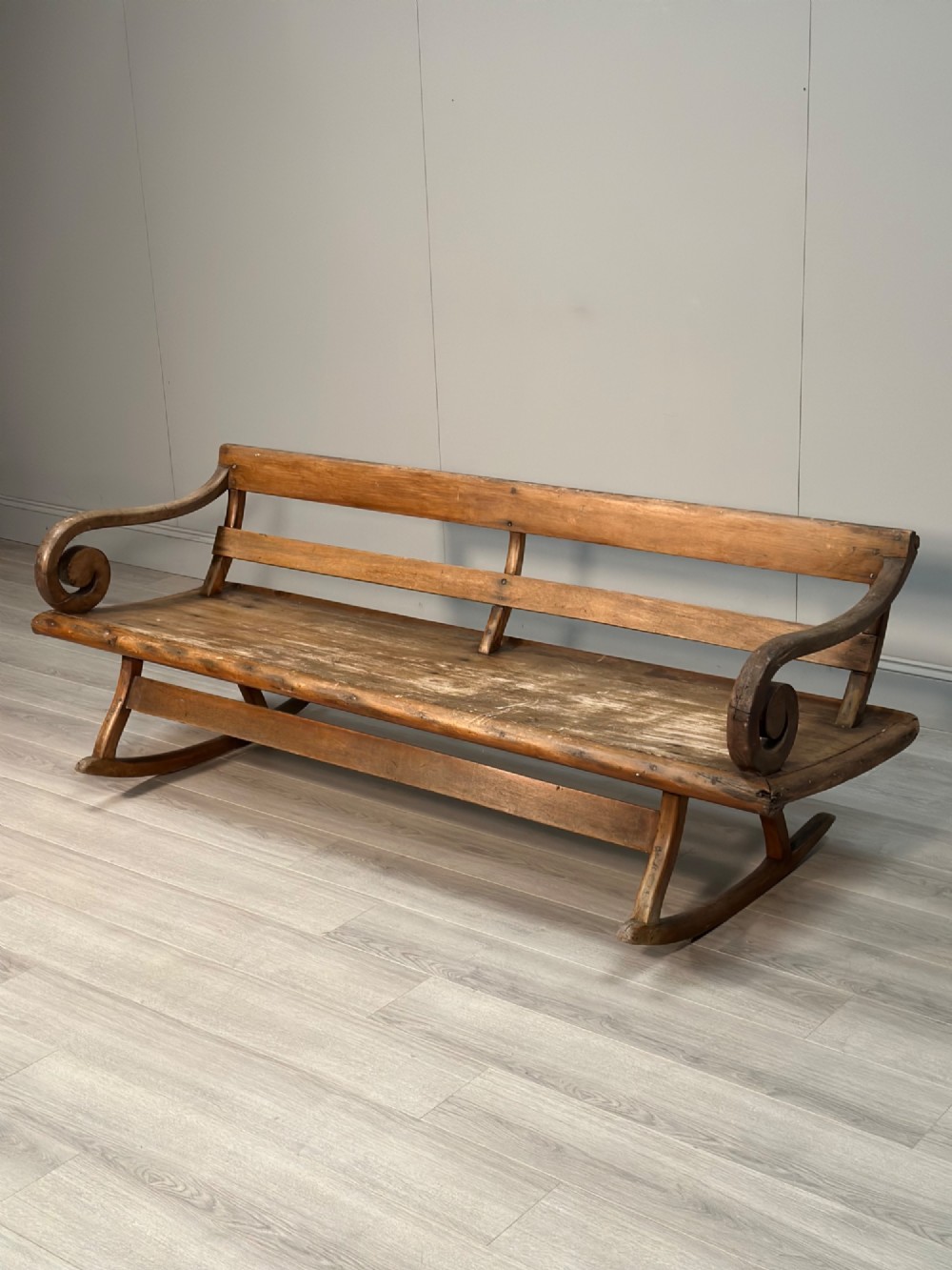 A long antique wooden rocking settle featuring open back slats, curved scroll armrests, and a solid plank seat resting on wooden rocking runners. View IX