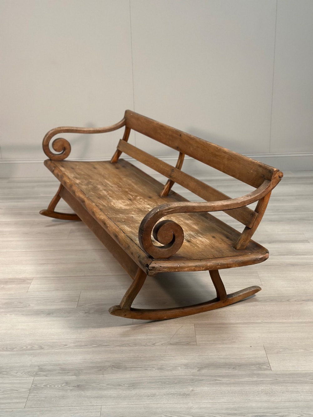 A long antique wooden rocking settle featuring open back slats, curved scroll armrests, and a solid plank seat resting on wooden rocking runners. View X