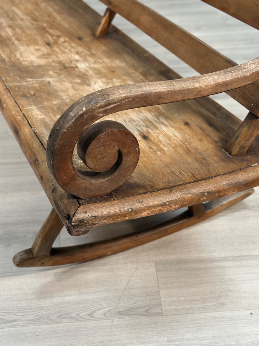 A long antique wooden rocking settle featuring open back slats, curved scroll armrests, and a solid plank seat resting on wooden rocking runners. View XI