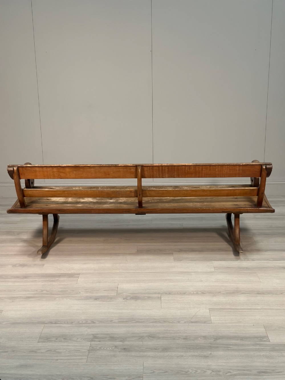 A long antique wooden rocking settle featuring open back slats, curved scroll armrests, and a solid plank seat resting on wooden rocking runners. View XIV