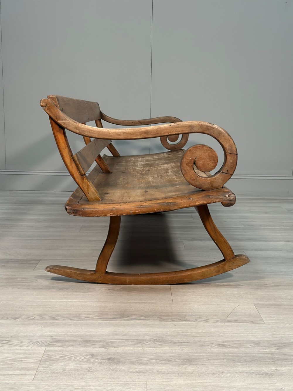A long antique wooden rocking settle featuring open back slats, curved scroll armrests, and a solid plank seat resting on wooden rocking runners. View XV