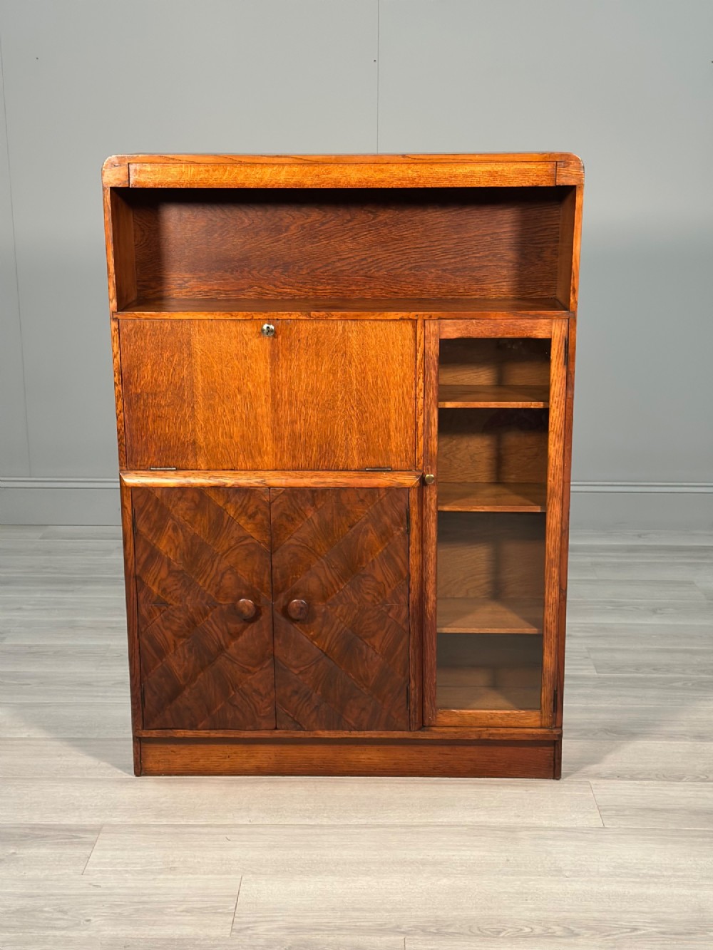 An Art Deco oak bureau bookcase featuring an open top shelf, drop-down writing flap, a glazed side cabinet, and lower cupboards with geometric diamond veneer.