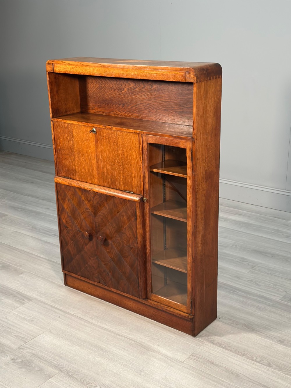 An Art Deco oak bureau bookcase featuring an open top shelf, drop-down writing flap, a glazed side cabinet, and lower cupboards with geometric diamond veneer. View II
