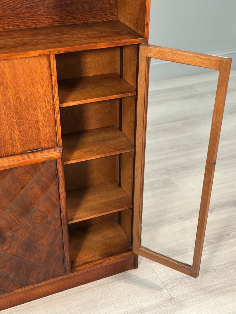 An Art Deco oak bureau bookcase featuring an open top shelf, drop-down writing flap, a glazed side cabinet, and lower cupboards with geometric diamond veneer. View VII