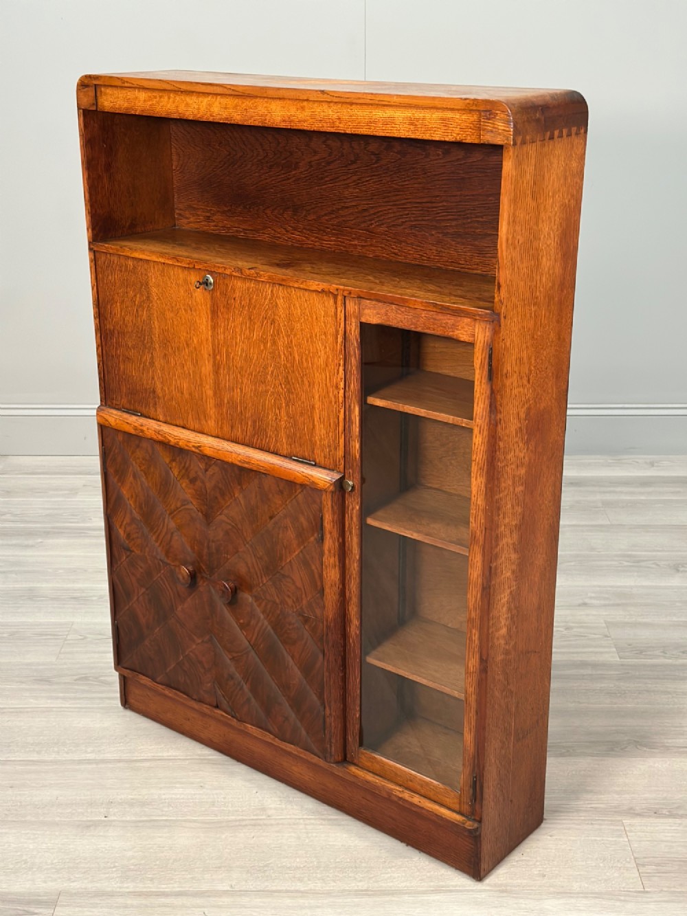 An Art Deco oak bureau bookcase featuring an open top shelf, drop-down writing flap, a glazed side cabinet, and lower cupboards with geometric diamond veneer. View VIII