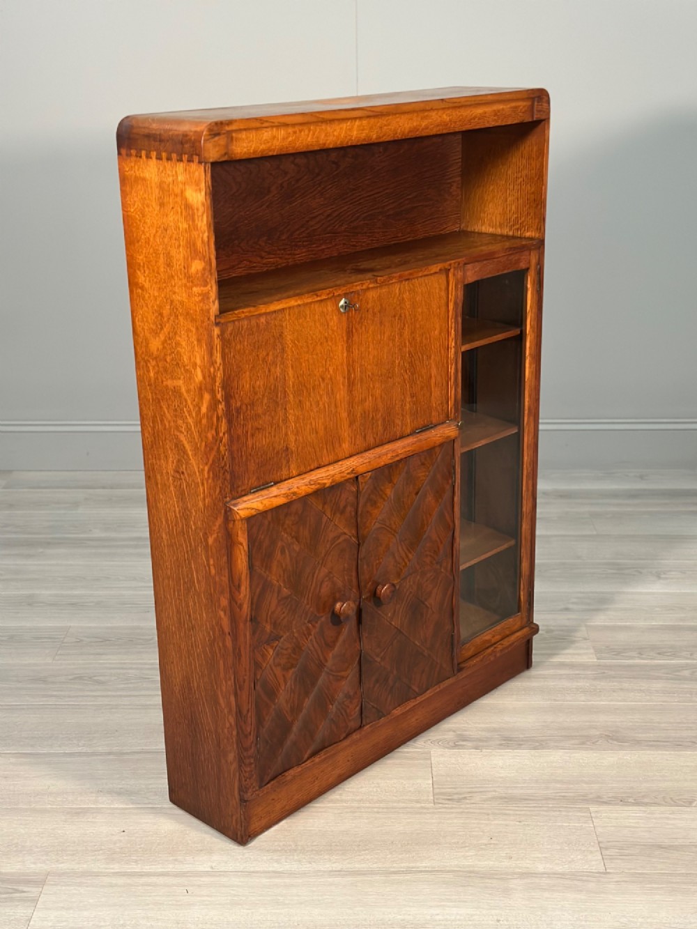 An Art Deco oak bureau bookcase featuring an open top shelf, drop-down writing flap, a glazed side cabinet, and lower cupboards with geometric diamond veneer. View XI