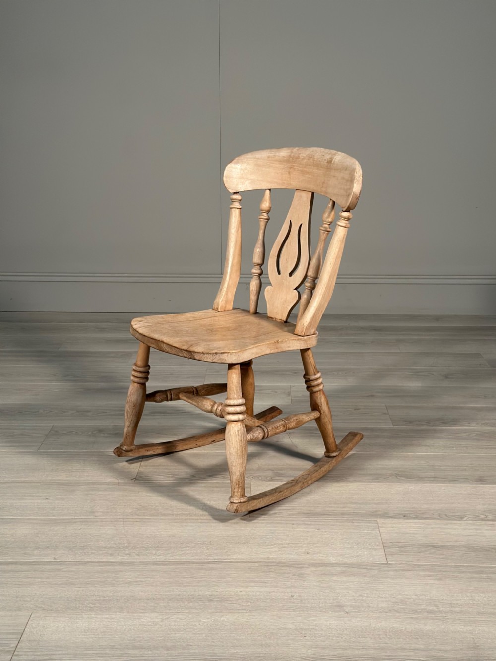 A small antique pale wood rocking chair featuring turned spindles, a pierced central back splat, and curved wooden rockers.