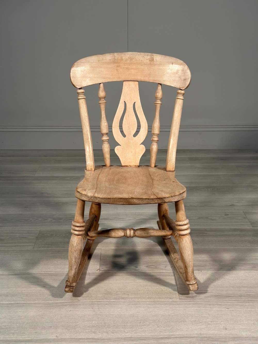 A small antique pale wood rocking chair featuring turned spindles, a pierced central back splat, and curved wooden rockers. View II