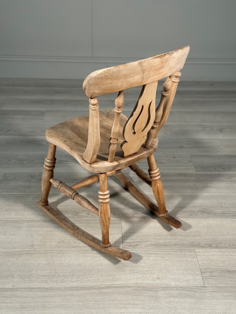 A small antique pale wood rocking chair featuring turned spindles, a pierced central back splat, and curved wooden rockers. View V