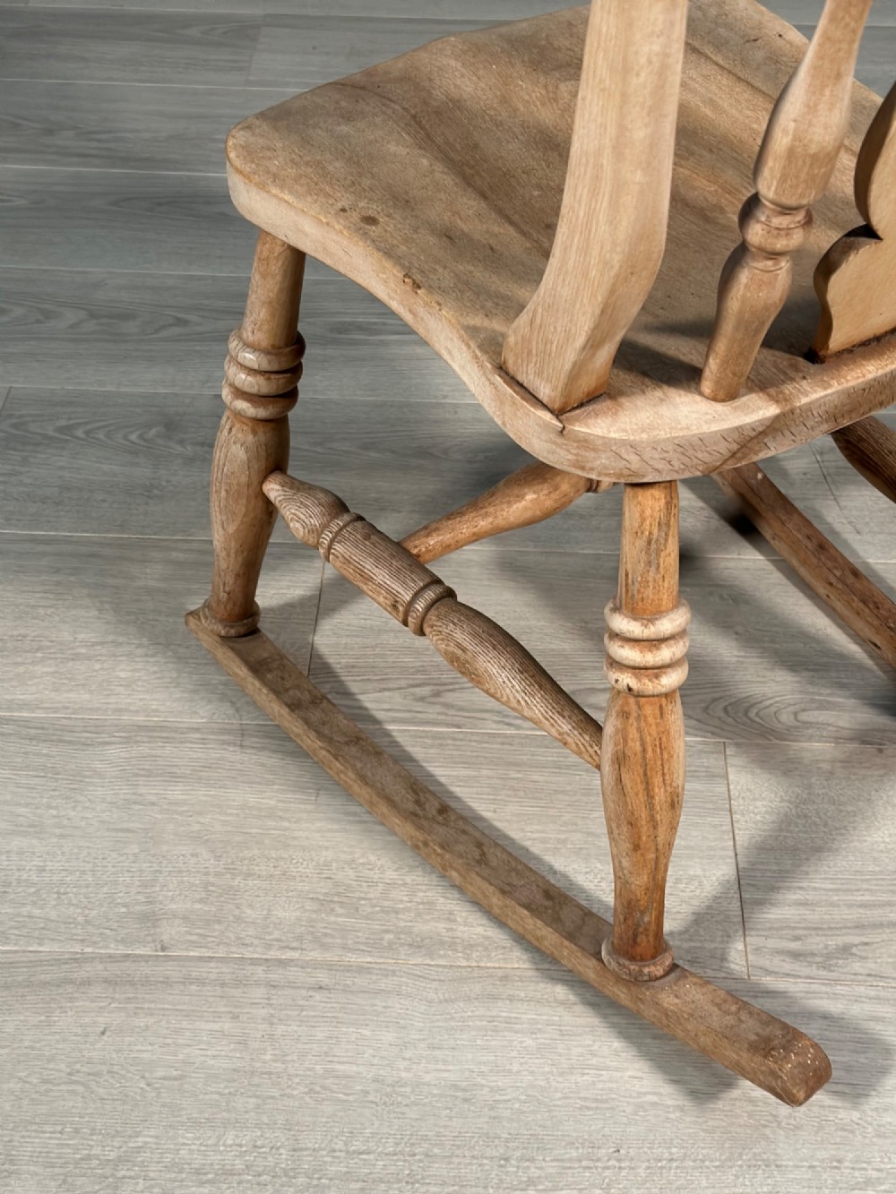 A small antique pale wood rocking chair featuring turned spindles, a pierced central back splat, and curved wooden rockers. View VI