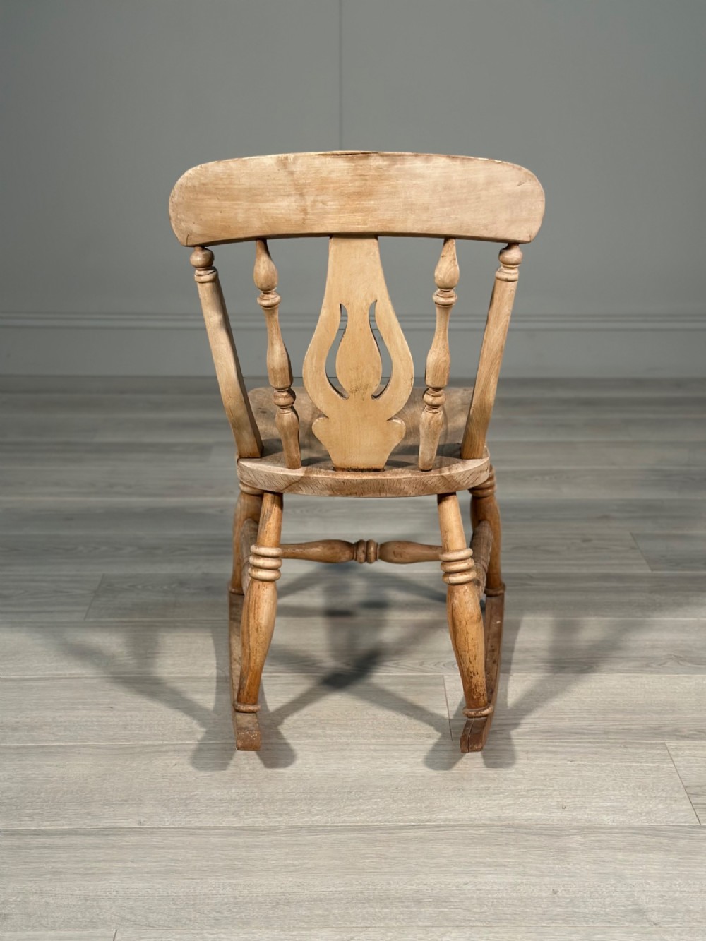 A small antique pale wood rocking chair featuring turned spindles, a pierced central back splat, and curved wooden rockers. View VIII