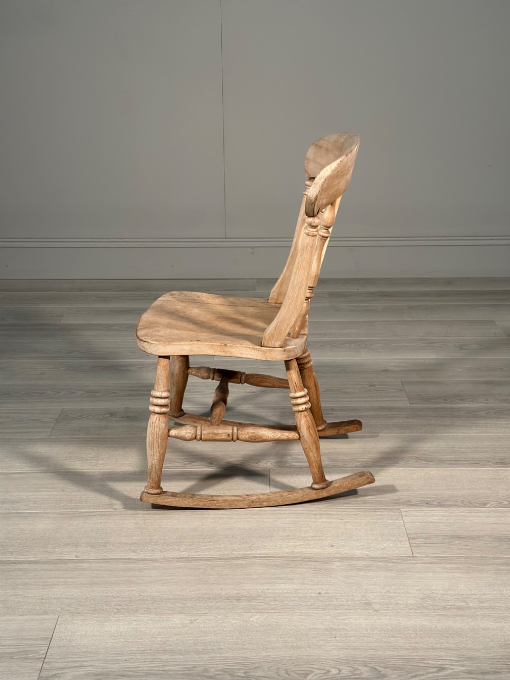 A small antique pale wood rocking chair featuring turned spindles, a pierced central back splat, and curved wooden rockers. View XI