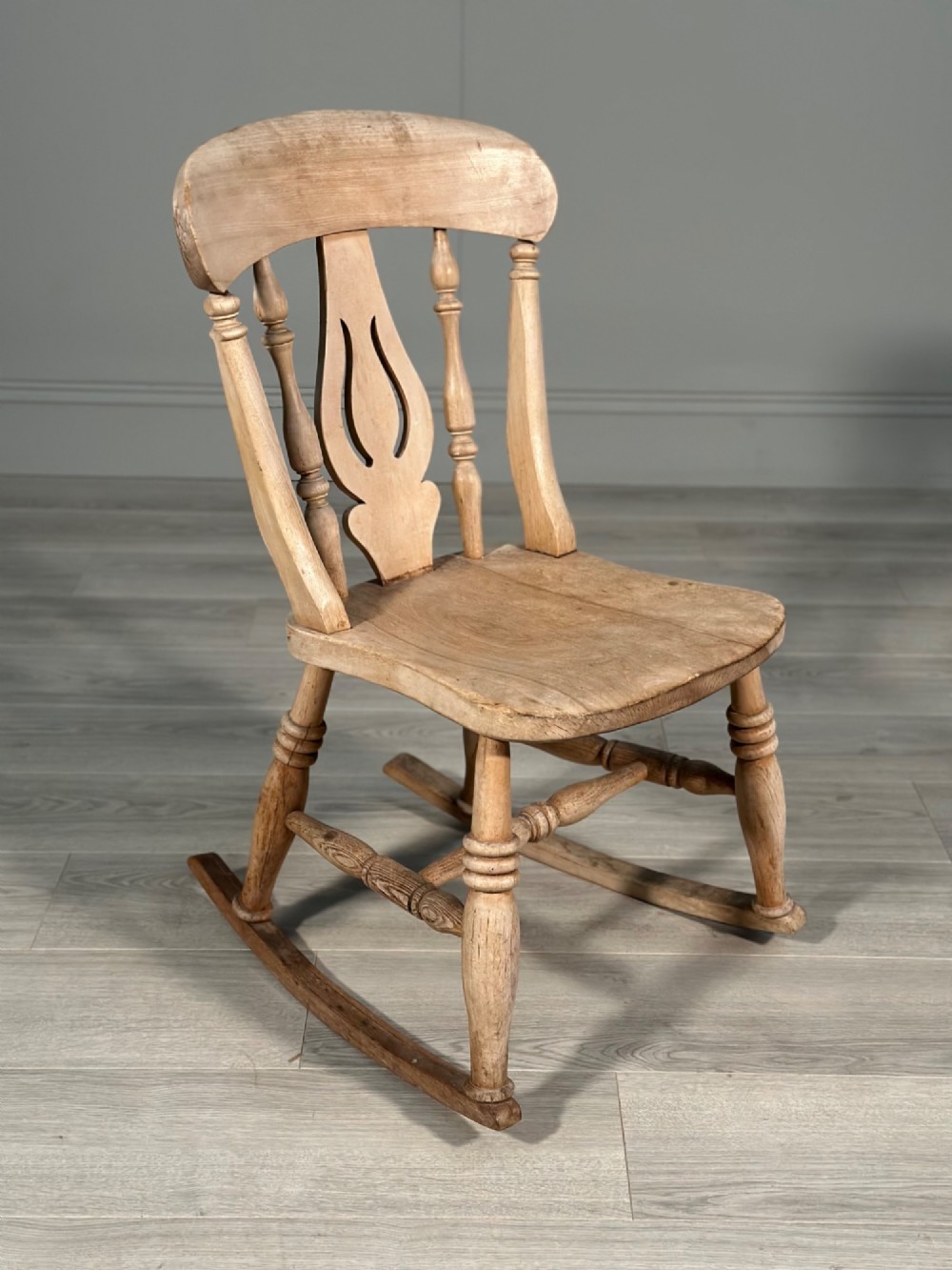 A small antique pale wood rocking chair featuring turned spindles, a pierced central back splat, and curved wooden rockers. View XII