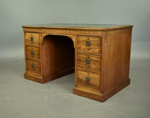 fig 1. A 19th-century Regency style mahogany pedestal desk featuring a leather writing top, carved wave frieze, six drawers with brass lion mask handles, and a central kneehole aperture.