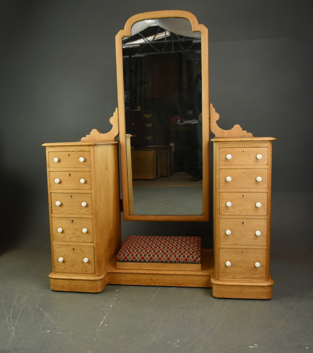 An antique Victorian pine dressing table featuring twin pedestals of five drawers each with white ceramic knobs, flanking a tall central arch-topped mirror and an upholstered footrest.