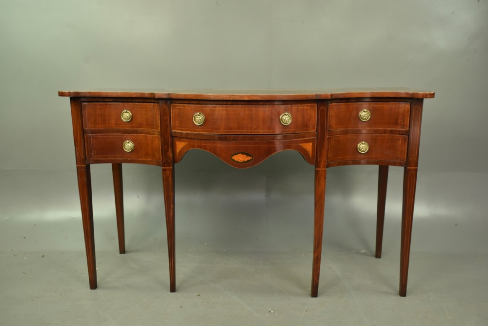 A Regency mahogany serpentine sideboard featuring curved drawer fronts, floral marquetry inlay, cast brass ring handles, and six tapered legs.