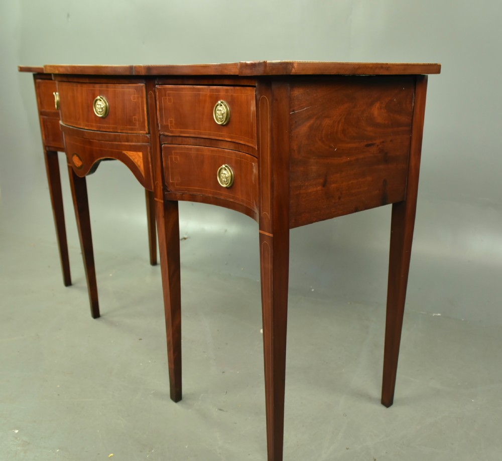 A Regency mahogany serpentine sideboard featuring curved drawer fronts, floral marquetry inlay, cast brass ring handles, and six tapered legs. View IX