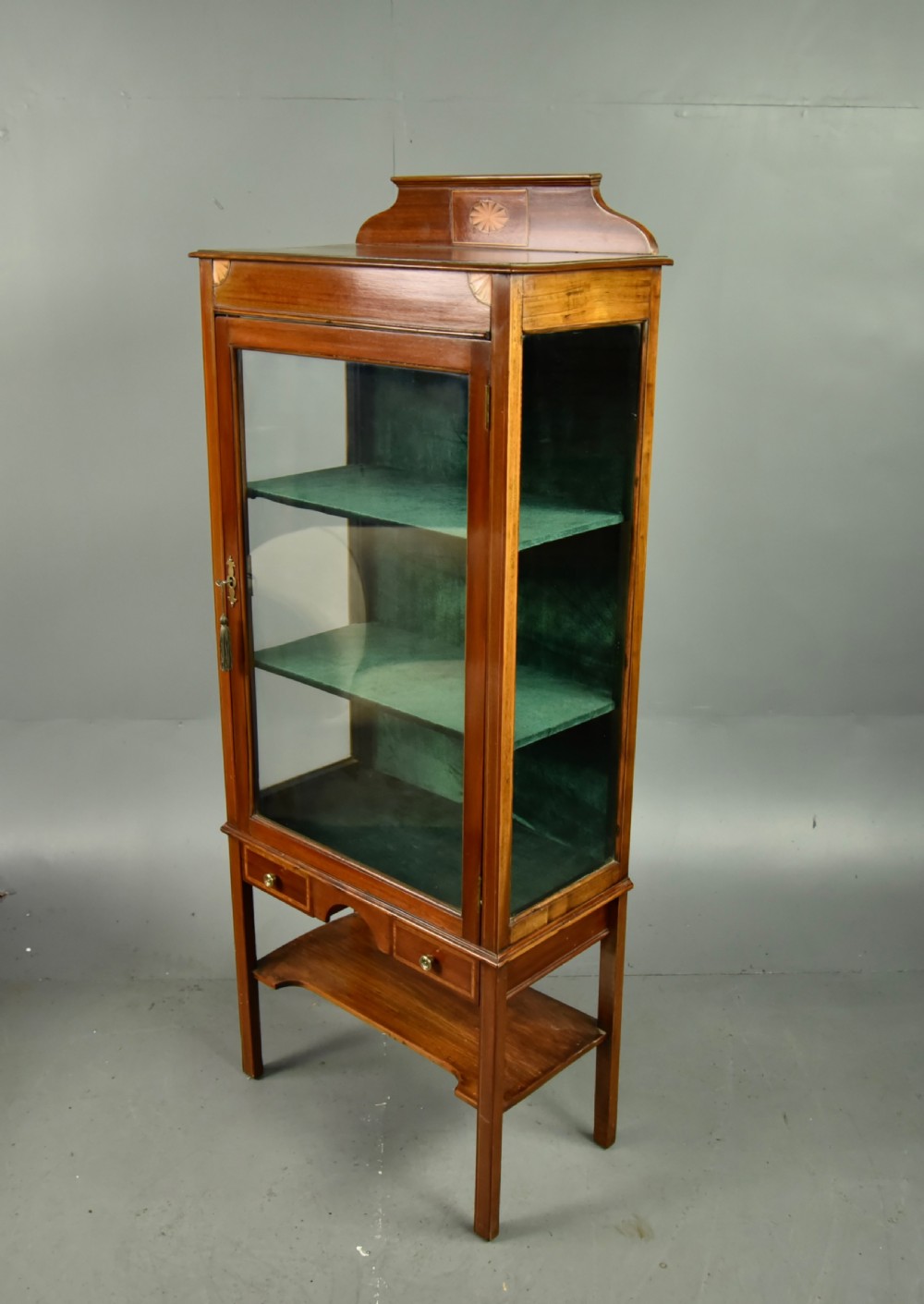 A narrow Edwardian mahogany display cabinet featuring a glazed door, green fabric-lined shelves, marquetry inlay on the upper pediment, and a lower shelf tier. View II