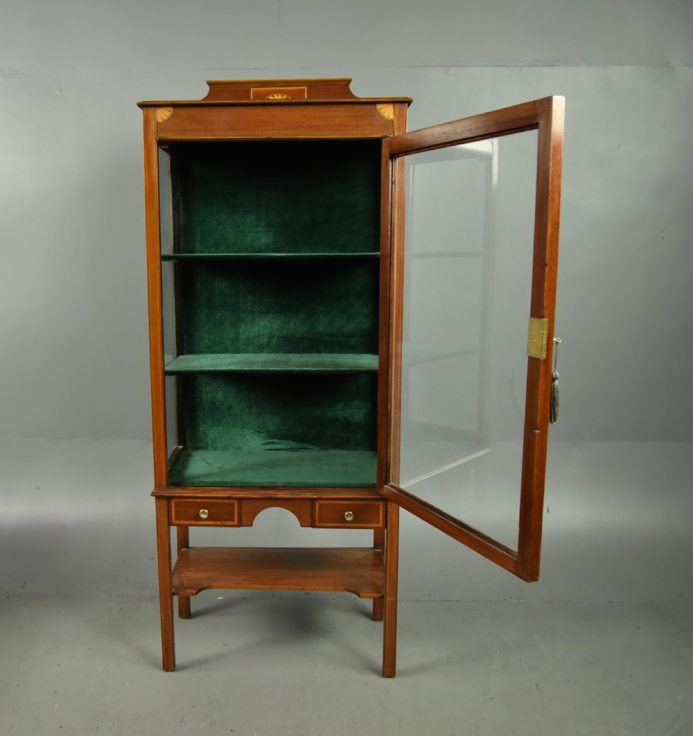 A narrow Edwardian mahogany display cabinet featuring a glazed door, green fabric-lined shelves, marquetry inlay on the upper pediment, and a lower shelf tier. View III
