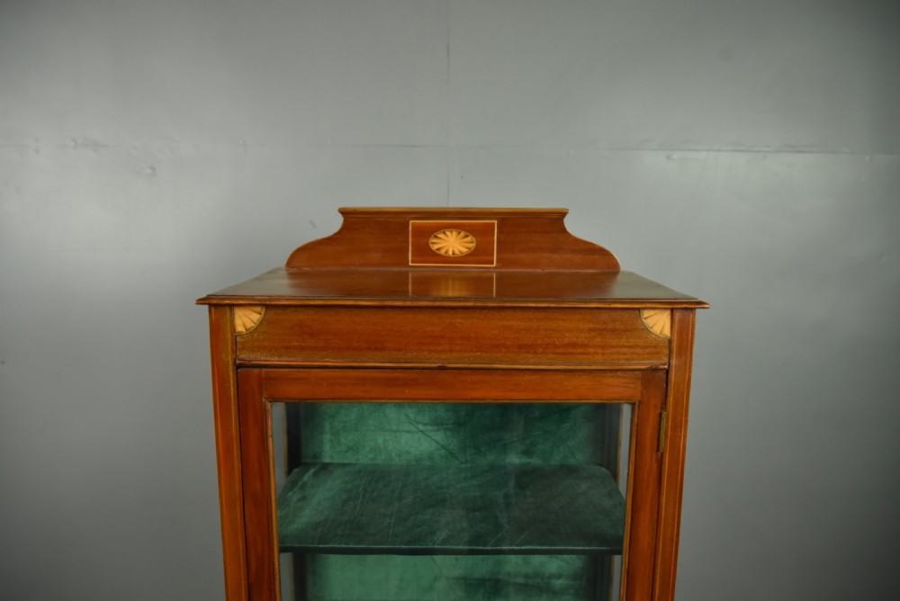 A narrow Edwardian mahogany display cabinet featuring a glazed door, green fabric-lined shelves, marquetry inlay on the upper pediment, and a lower shelf tier. View IV