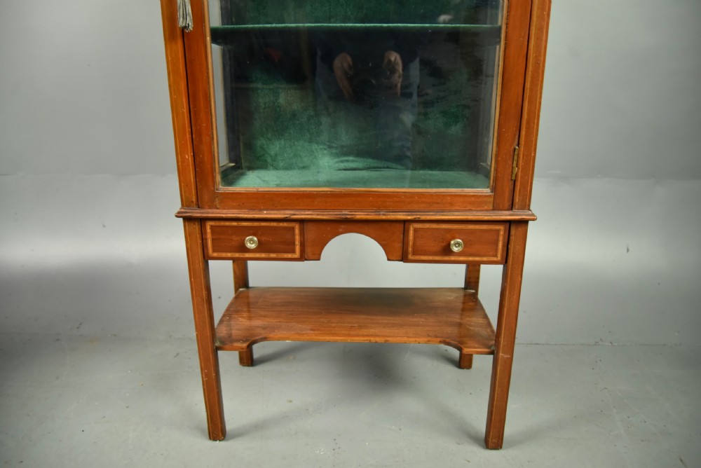 A narrow Edwardian mahogany display cabinet featuring a glazed door, green fabric-lined shelves, marquetry inlay on the upper pediment, and a lower shelf tier. View V