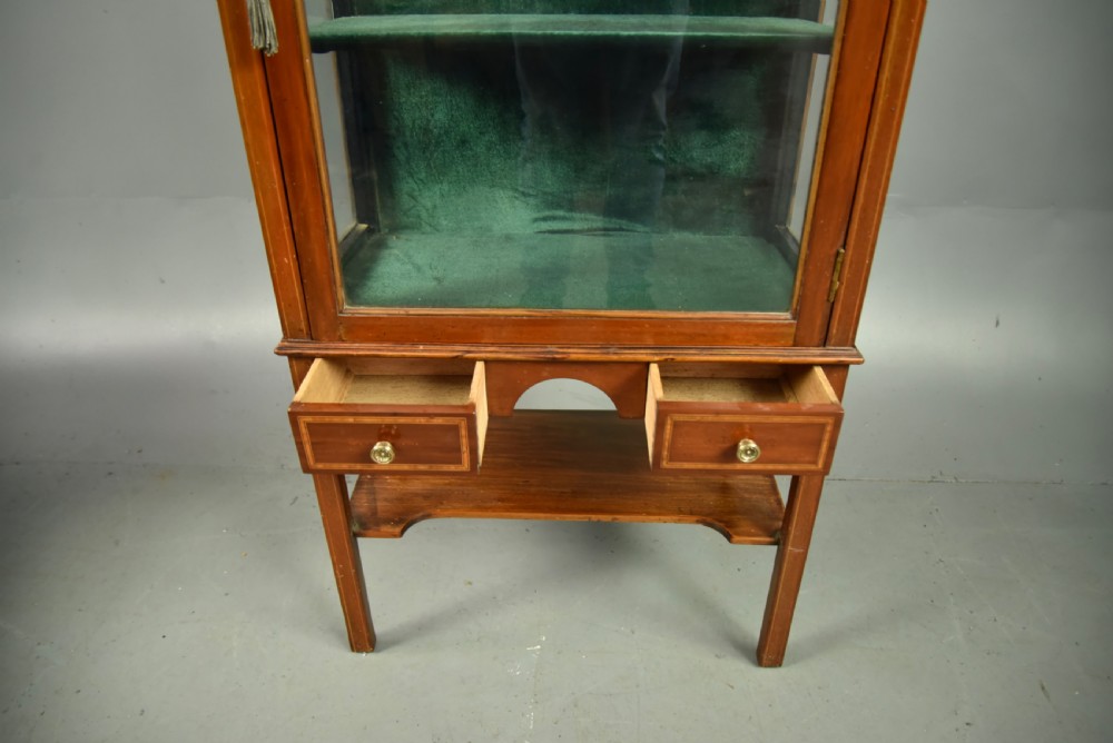 A narrow Edwardian mahogany display cabinet featuring a glazed door, green fabric-lined shelves, marquetry inlay on the upper pediment, and a lower shelf tier. View VI