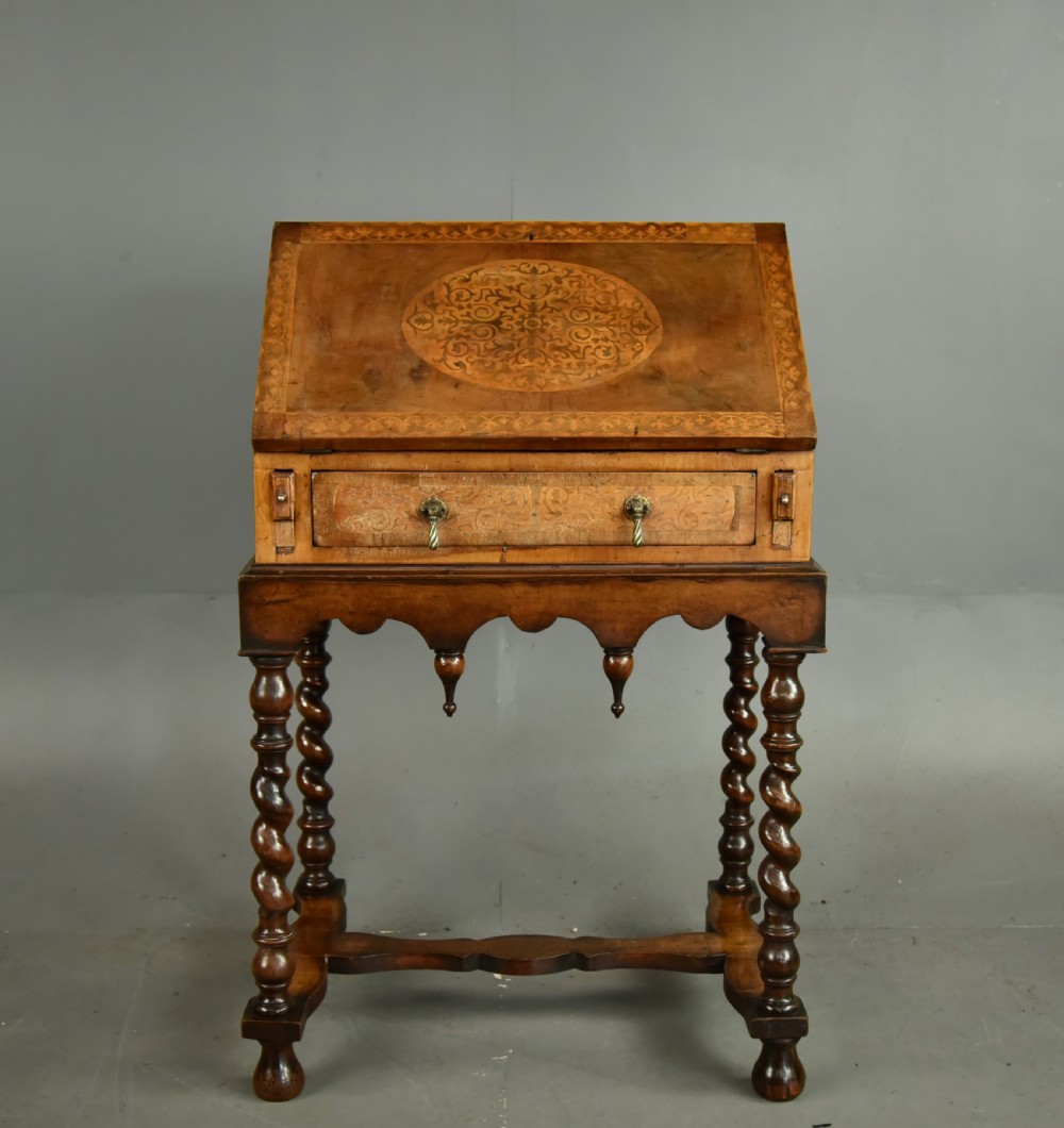 A small walnut bureau featuring an oval floral marquetry panel on the sloped front, set above a drawer and supported by four barley twist legs.