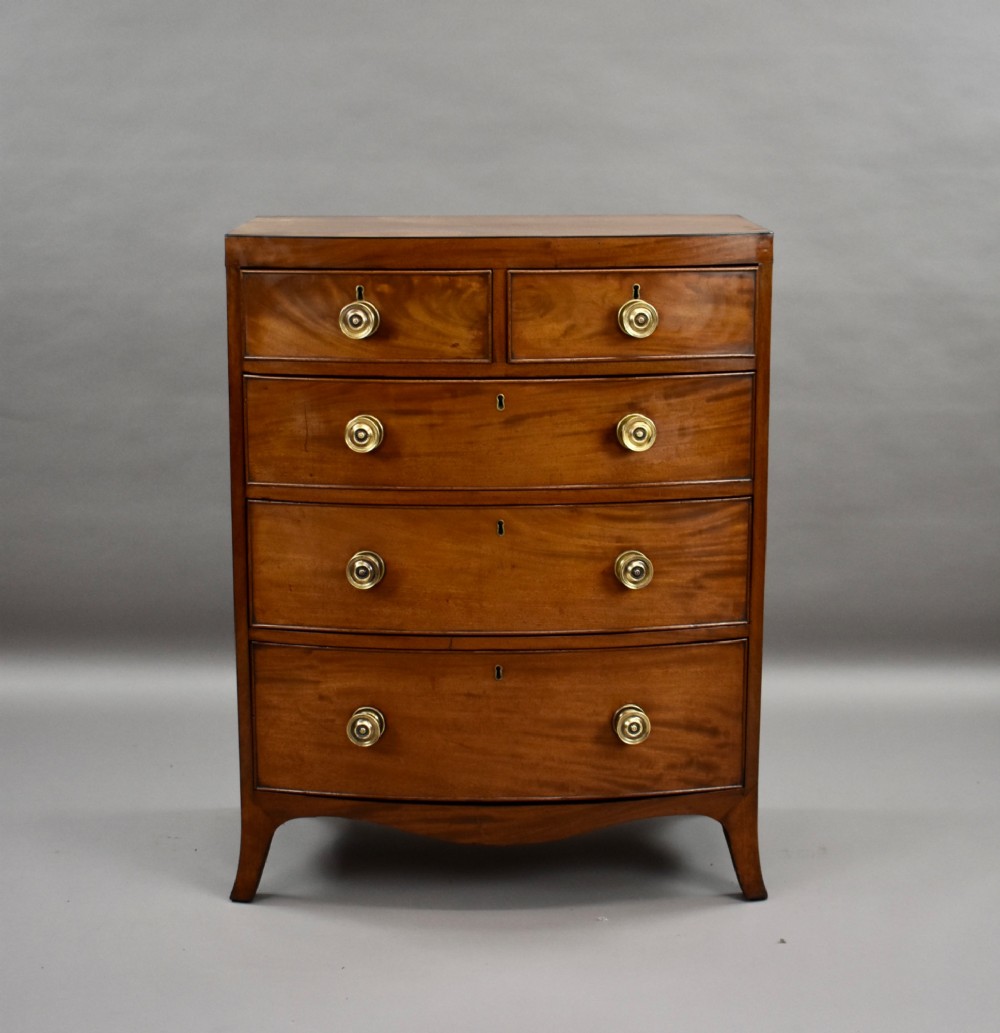 A narrow Regency mahogany bowfront chest featuring two short over three long drawers with turned brass handles and splayed bracket feet. View II