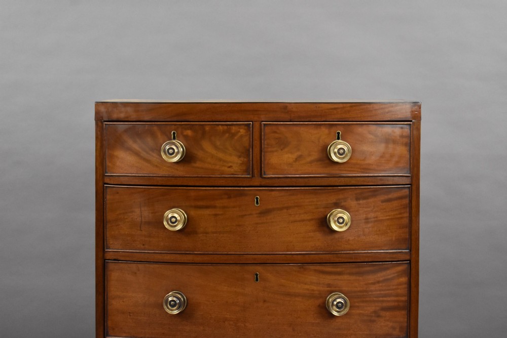 A narrow Regency mahogany bowfront chest featuring two short over three long drawers with turned brass handles and splayed bracket feet. View III