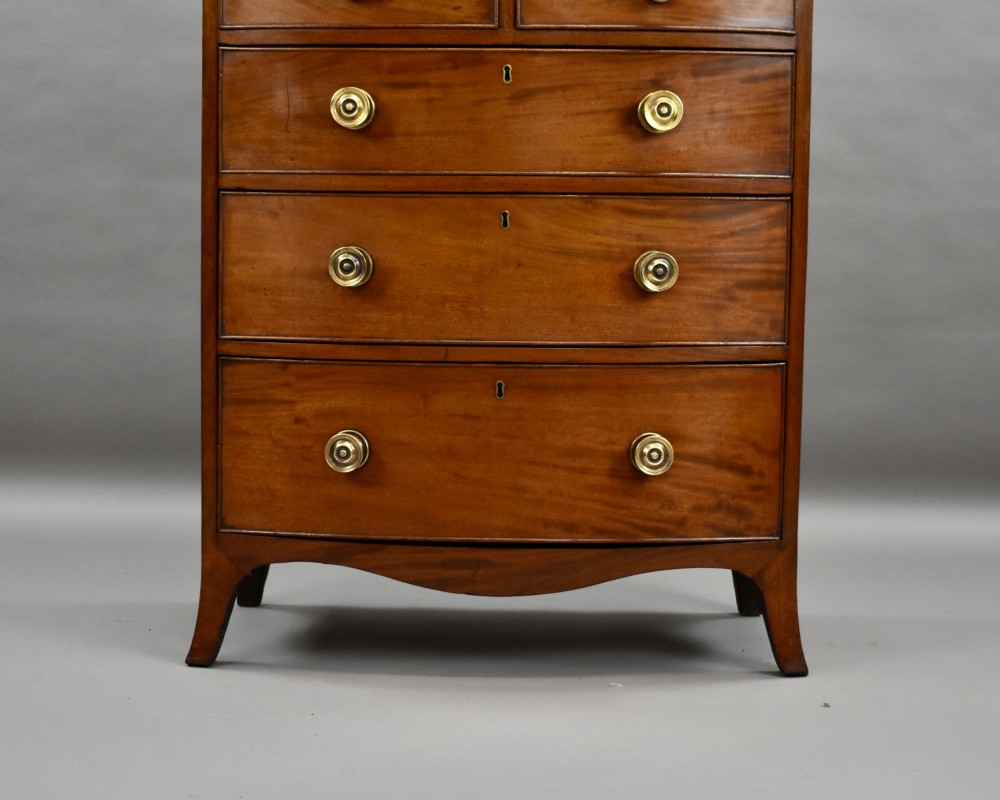 A narrow Regency mahogany bowfront chest featuring two short over three long drawers with turned brass handles and splayed bracket feet. View IV