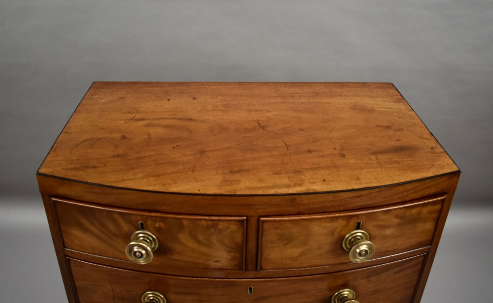 A narrow Regency mahogany bowfront chest featuring two short over three long drawers with turned brass handles and splayed bracket feet. View V