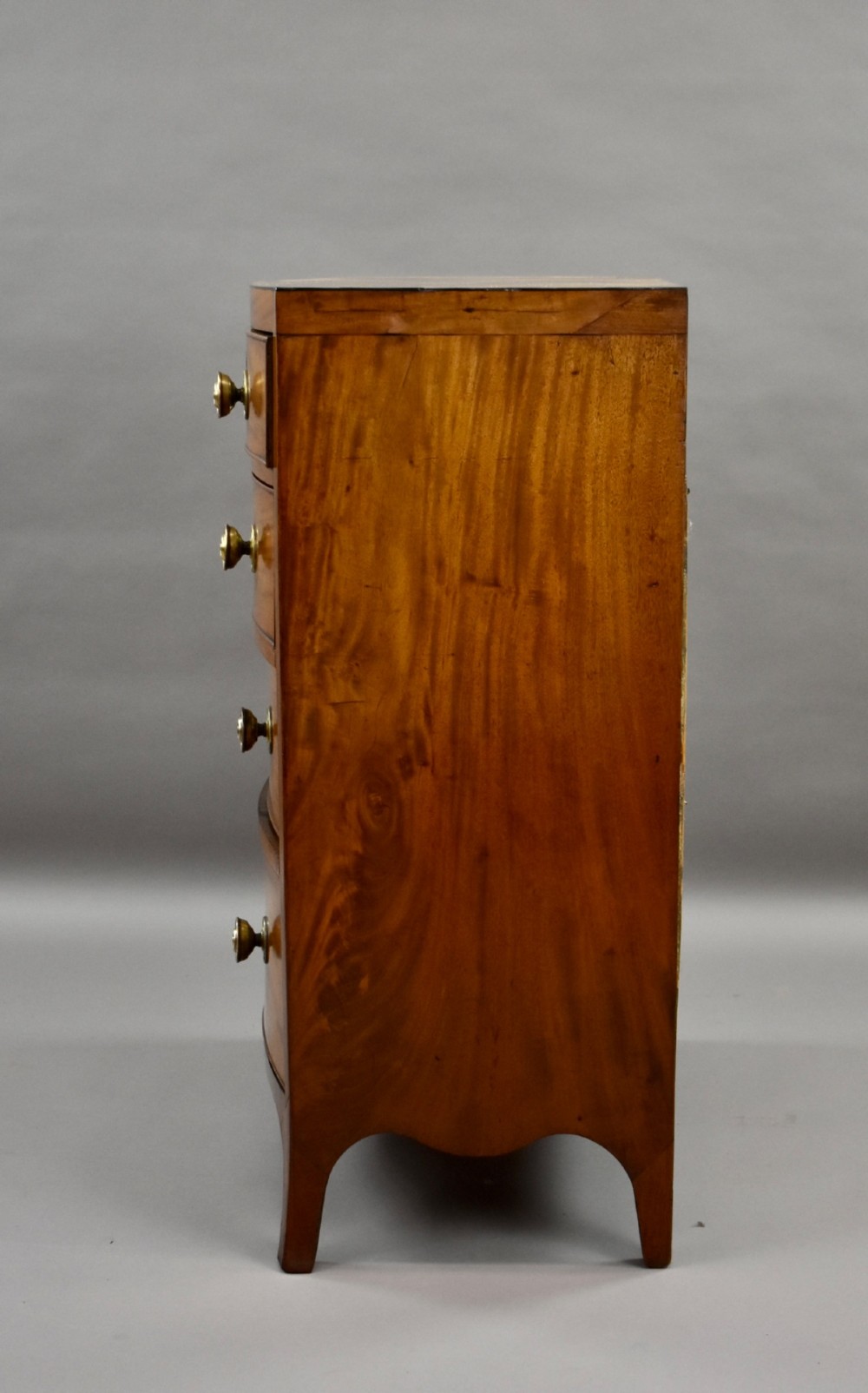 A narrow Regency mahogany bowfront chest featuring two short over three long drawers with turned brass handles and splayed bracket feet. View VI
