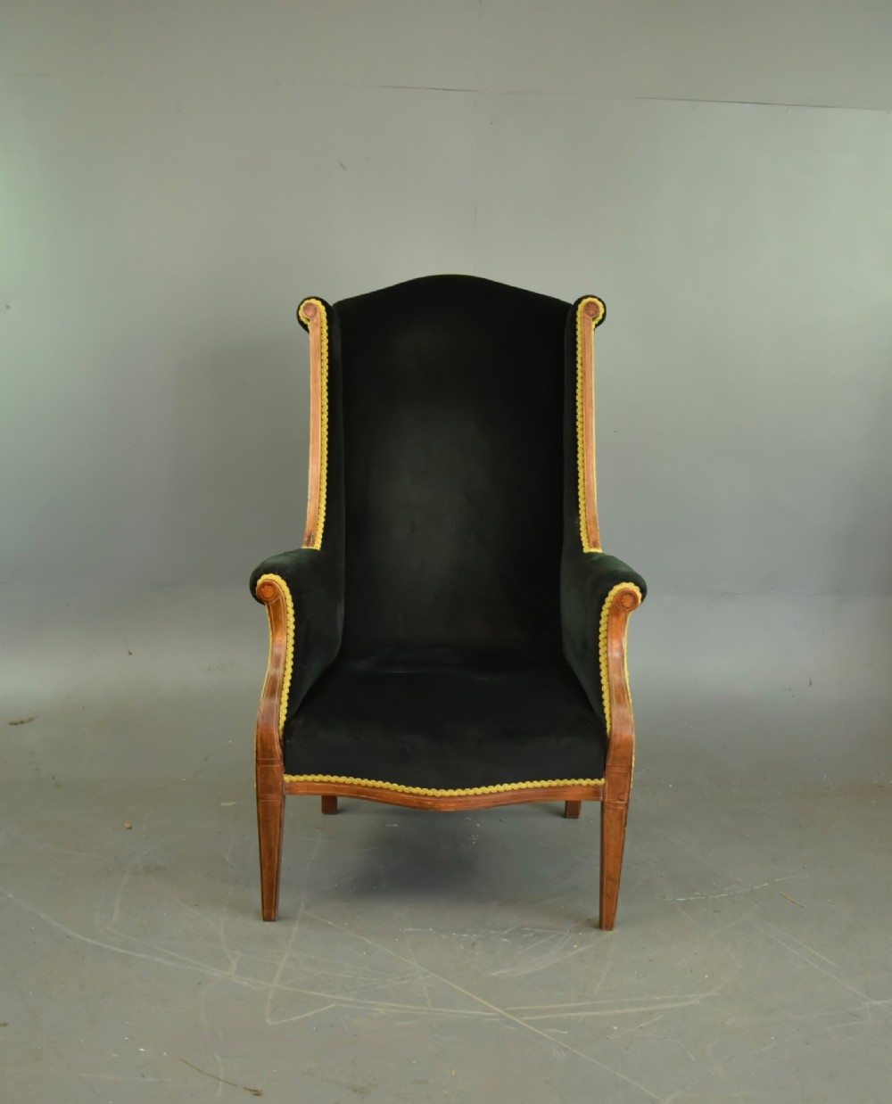 An Edwardian mahogany wing armchair upholstered in dark green velvet with contrasting decorative gimp trim and tapered front legs. View II