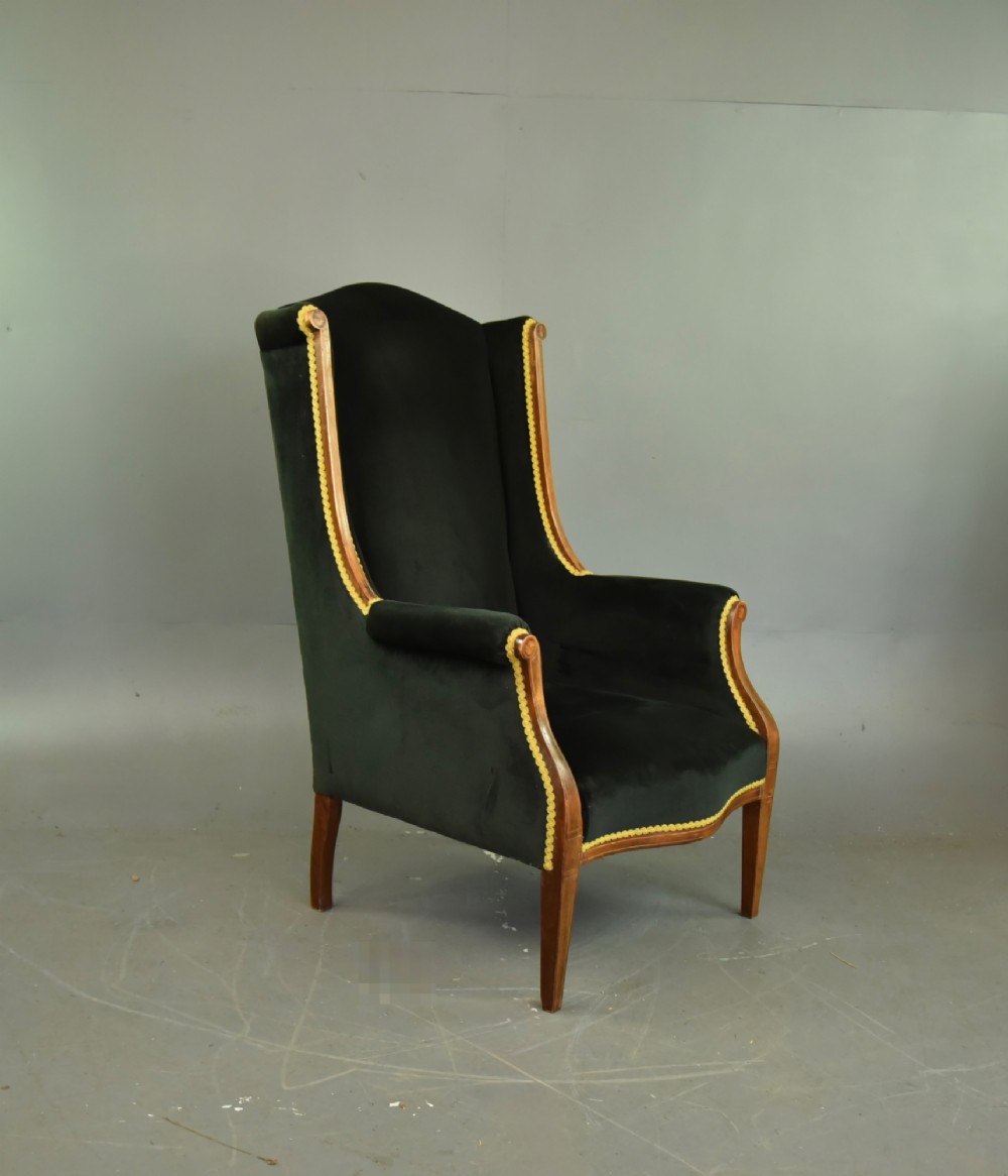 An Edwardian mahogany wing armchair upholstered in dark green velvet with contrasting decorative gimp trim and tapered front legs. View III
