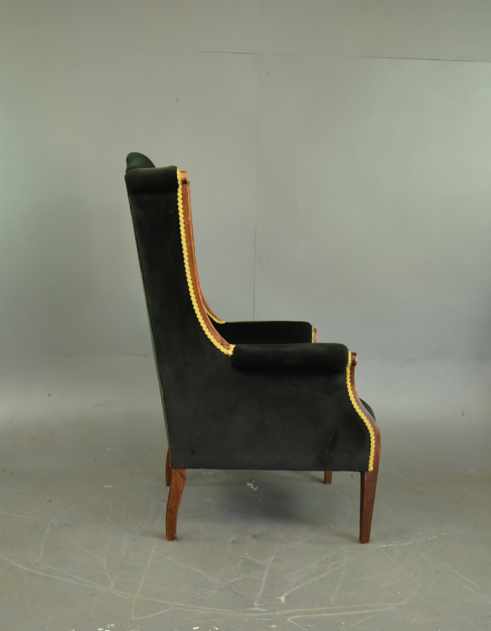 An Edwardian mahogany wing armchair upholstered in dark green velvet with contrasting decorative gimp trim and tapered front legs. View IV