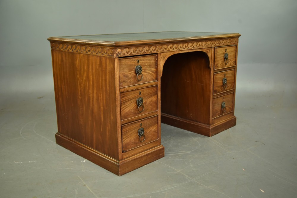 A 19th-century Regency style mahogany pedestal desk featuring a leather writing top, carved wave frieze, six drawers with brass lion mask handles, and a central kneehole aperture. View VII