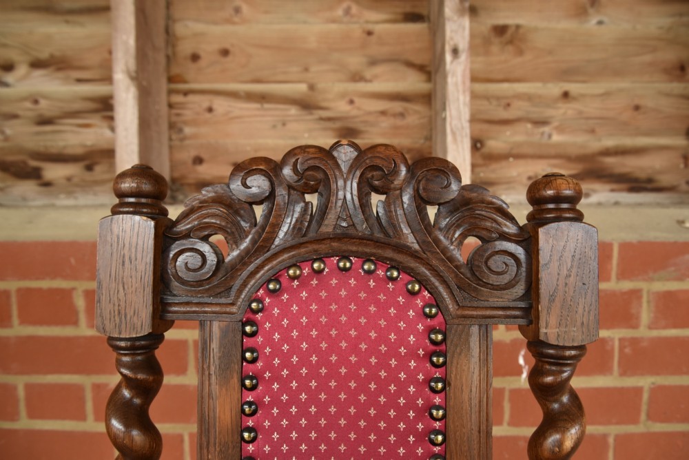 A set of eight carved oak dining chairs featuring barley twist supports, high carved crests, and red upholstered seats and backrests with brass stud detailing. View X