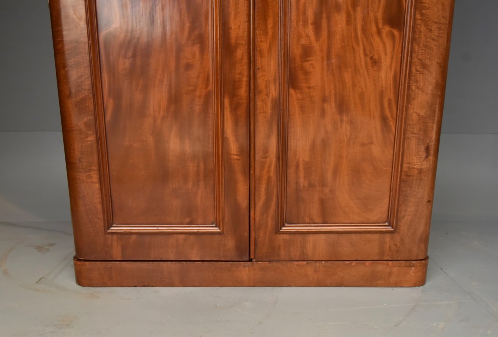 A Victorian flame mahogany double wardrobe featuring two arched panel doors, a moulded top cornice, and a grounded plinth base. View V